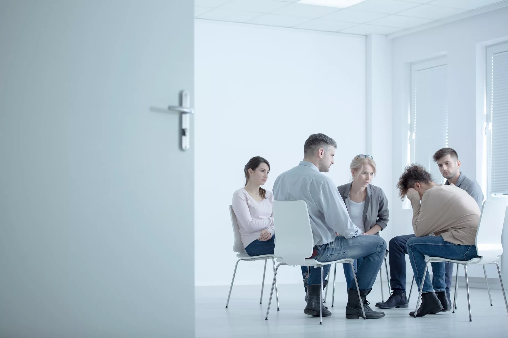 stark stock photo with much white space showing a woman crying in a small seated group at a rehab center with an open door in the foreground