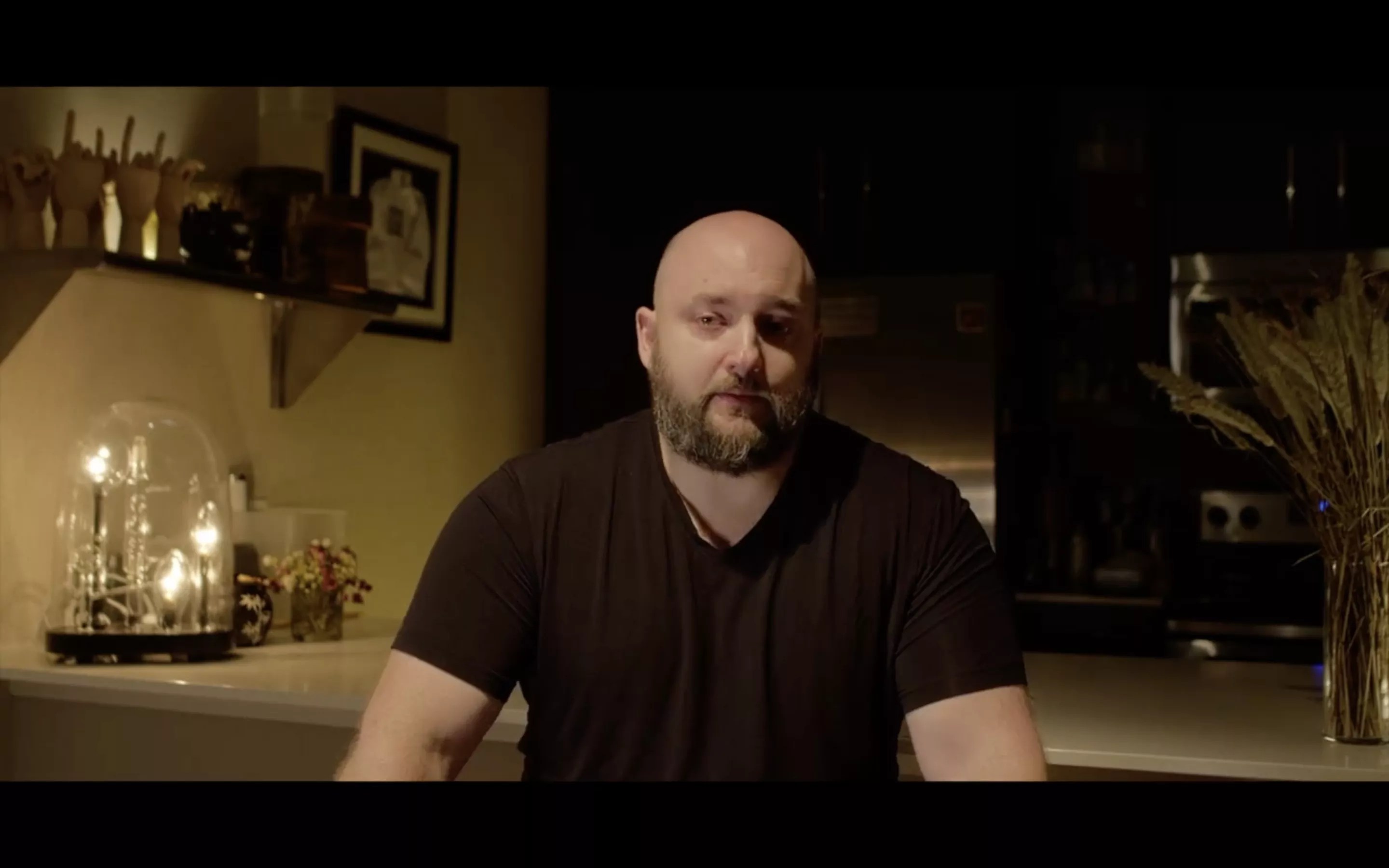 screenshot of a man with a shaved head and a full beard, seated at a kitchen counter