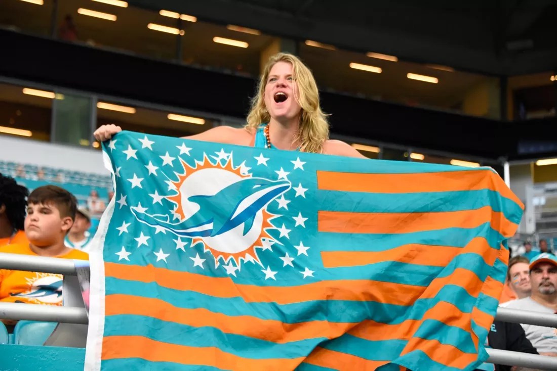 A blonde woman holds a Miami Dolphins flag with teal and orange stripes on it